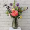 Full Bodied Hero White Wall The Show Stopper bouquet — an abundant, large-scale arrangement of vibrant seasonal blooms in rich tones, overflowing from a black glass vase against a pale brick backdrop.