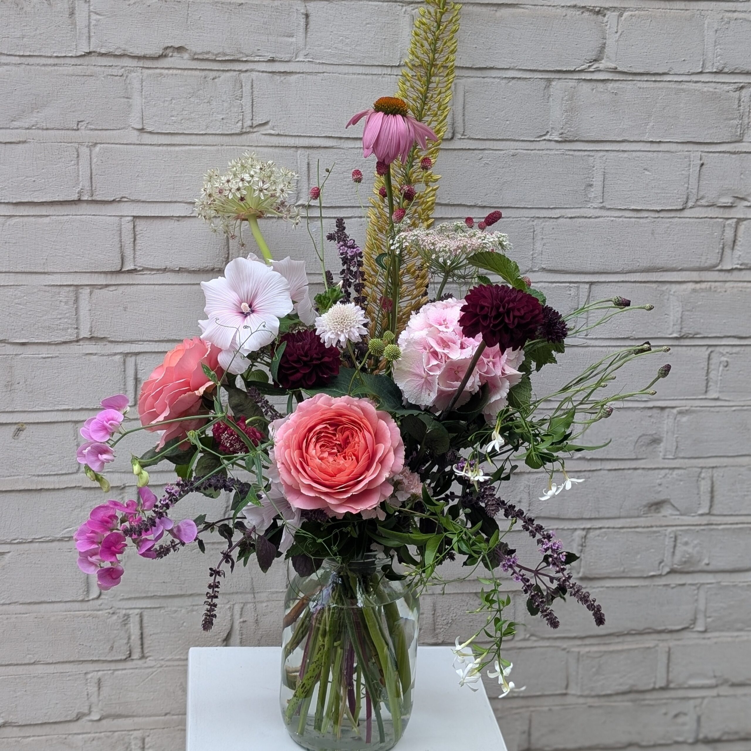 A summer bouquet in a clear jar, with coral garden roses, sweet peas, deep burgundy dahlias, and soft pink hollyhocks, styled in front of a pale brick wall
