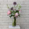 The Mother of Pearl Bouquet — an elegant arrangement of blush, cream, and shell-pink British blooms in a clear glass vase, sitting on a white plinth against a pale brick wall.