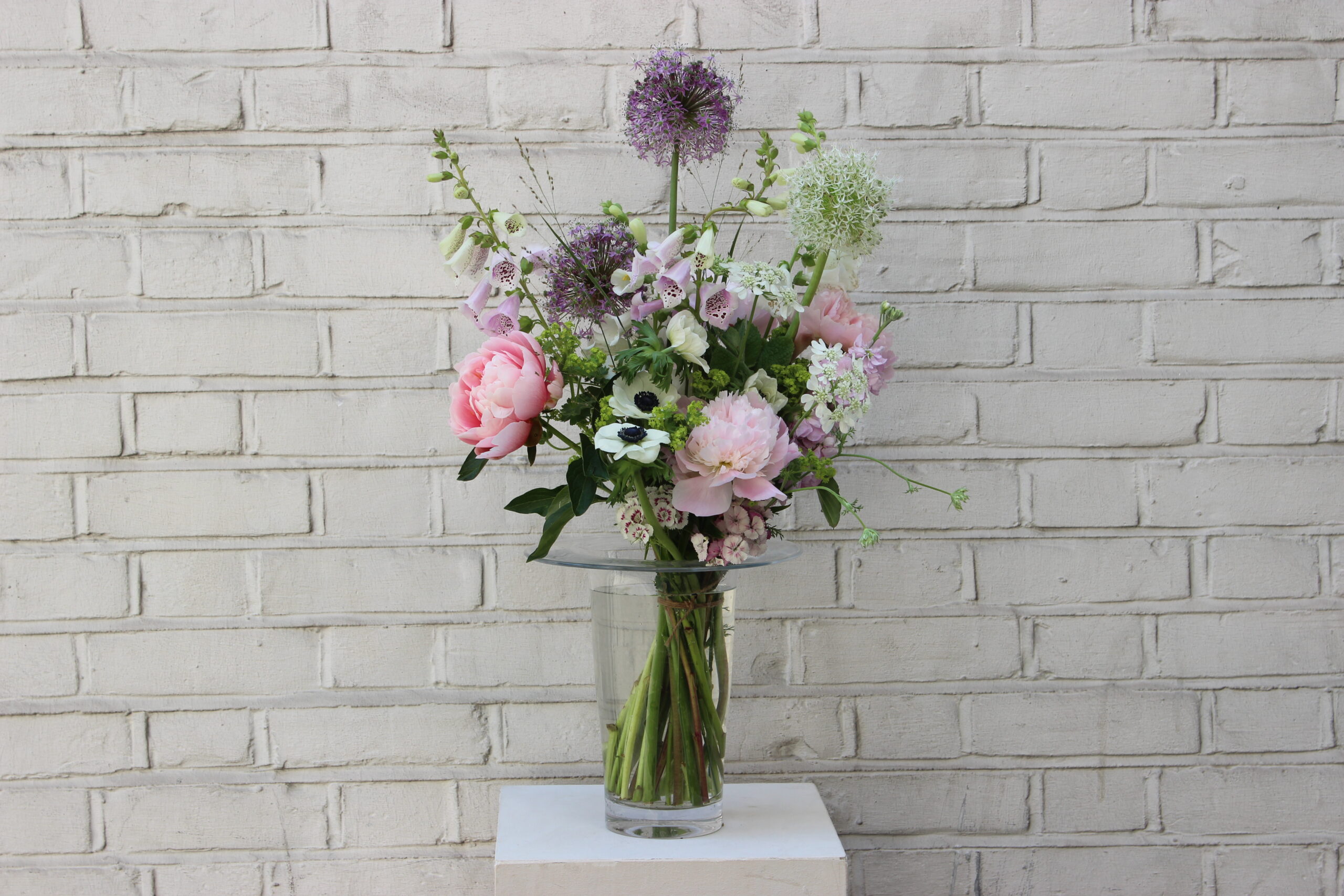 The Mother of Pearl Bouquet — an elegant arrangement of blush, cream, and shell-pink British blooms in a clear glass vase, sitting on a white plinth against a pale brick wall.