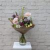 Summer Cordial Hero The Summer Cordial Bouquet by Stem and Stem, arranged in a clear glass jar against a pale brick wall. A warm, seasonal mix of British flowers including dahlias, cosmos, allium, and queen anne’s lace, wrapped in kraft paper.