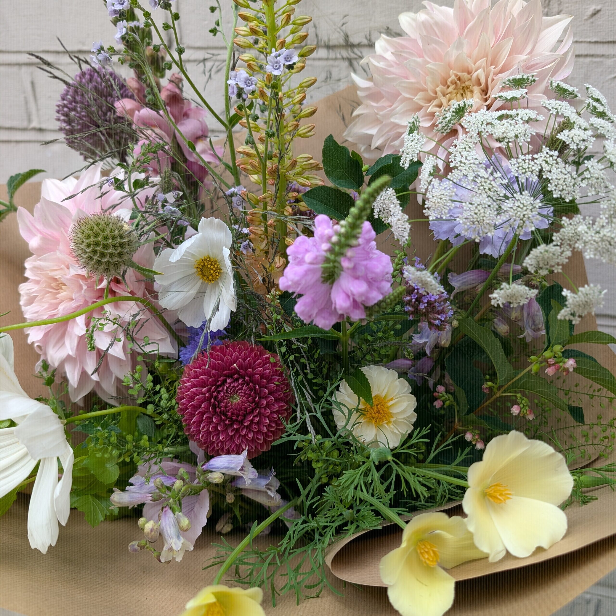 A soft, seasonal bouquet of British summer flowers in gentle tones of blush, lemon, lilac, and cream. Featuring cafe-au-lait dahlias, cosmos, scabious, pincushion flower, and delicate grasses, wrapped in brown paper.
